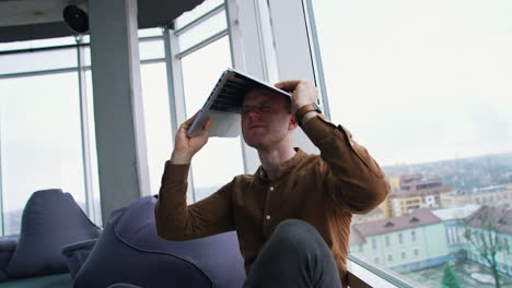 tired businessman with a laptop on his head. man is sitting in the office by the large window and holding a laptop over the head. problems in business.