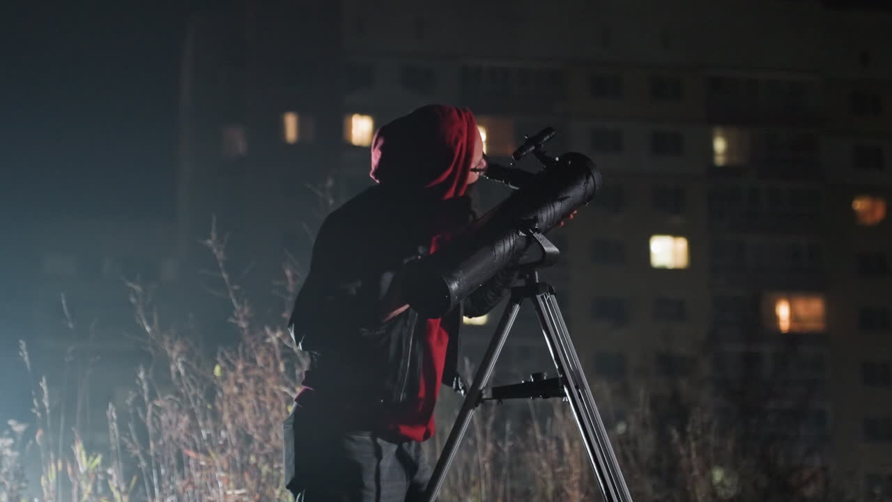 Persona che modifica un telescopio su un treppiede sotto le luci della città, calibratura e allineamento metodici con un notebook, finestre di appartamenti illuminate sullo sfondo, misurazione notturna precisa e impostazione dell'attrezzatura
