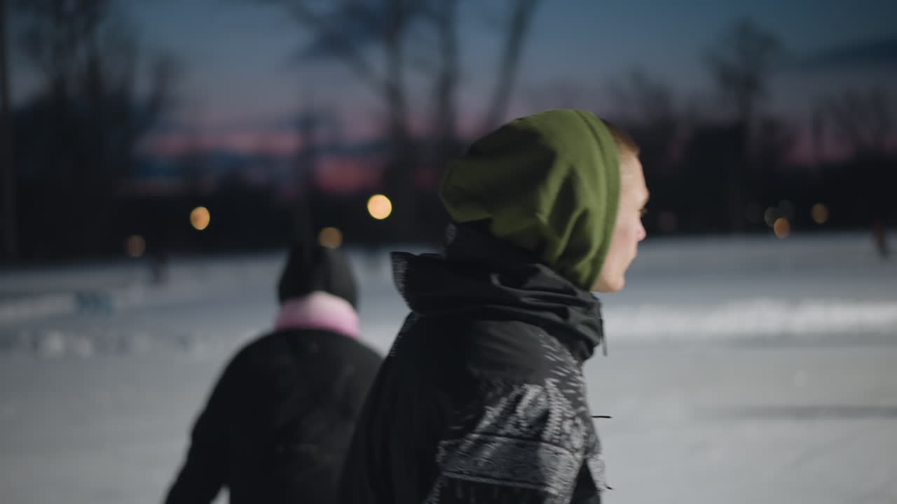 rear view student skating on outdoor ice rink at dusk wearing patterned jacket and green hoodie with blurred friends laughing and playing together creating joyful winter atmosphere
