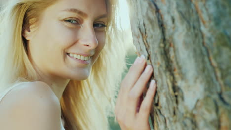 portrait of a beautiful young woman standing by the tree in the sun smiling looking at the camera co