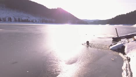 descending aerial view of man neck deep in frozen mountain lake with sunlight shining on icy surface