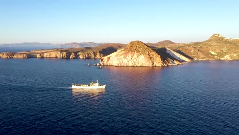 aerial drone shot of a large fishing boat cruising past the rocky cliff coastline during sunrise or sunset in milos greece