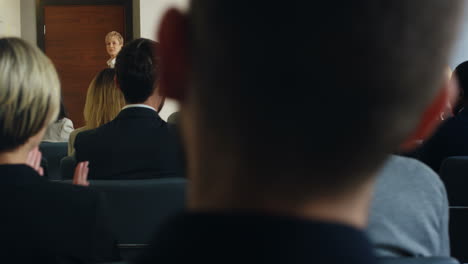 rear view of people sitting at a conference, while a businesswoman going into the conference room and coming up onto the podium