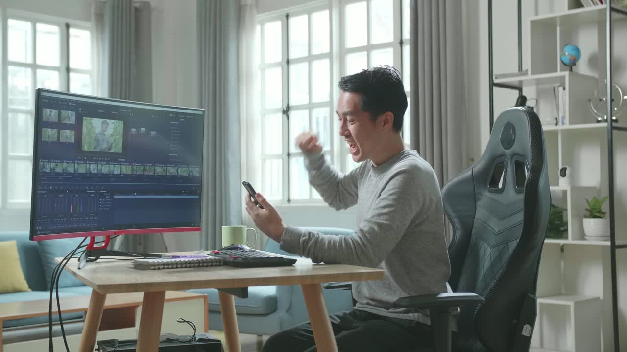 Asian Video Editor Man In Long Sleeved T-Shirt And Black Pants Looking At Mobile Phone And Being Happy Due To Success While Using Desktop Computer For Editing Video At Home.