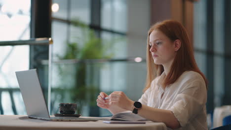 thoughtful-brooding-remote-working-red-haired-woman-sitting-infront-of-a-laptop-or-notebook-in-casual-outfit-on-her-work-desk-in-her-modern-airy-bright-living-room-home-office-with-many-windows