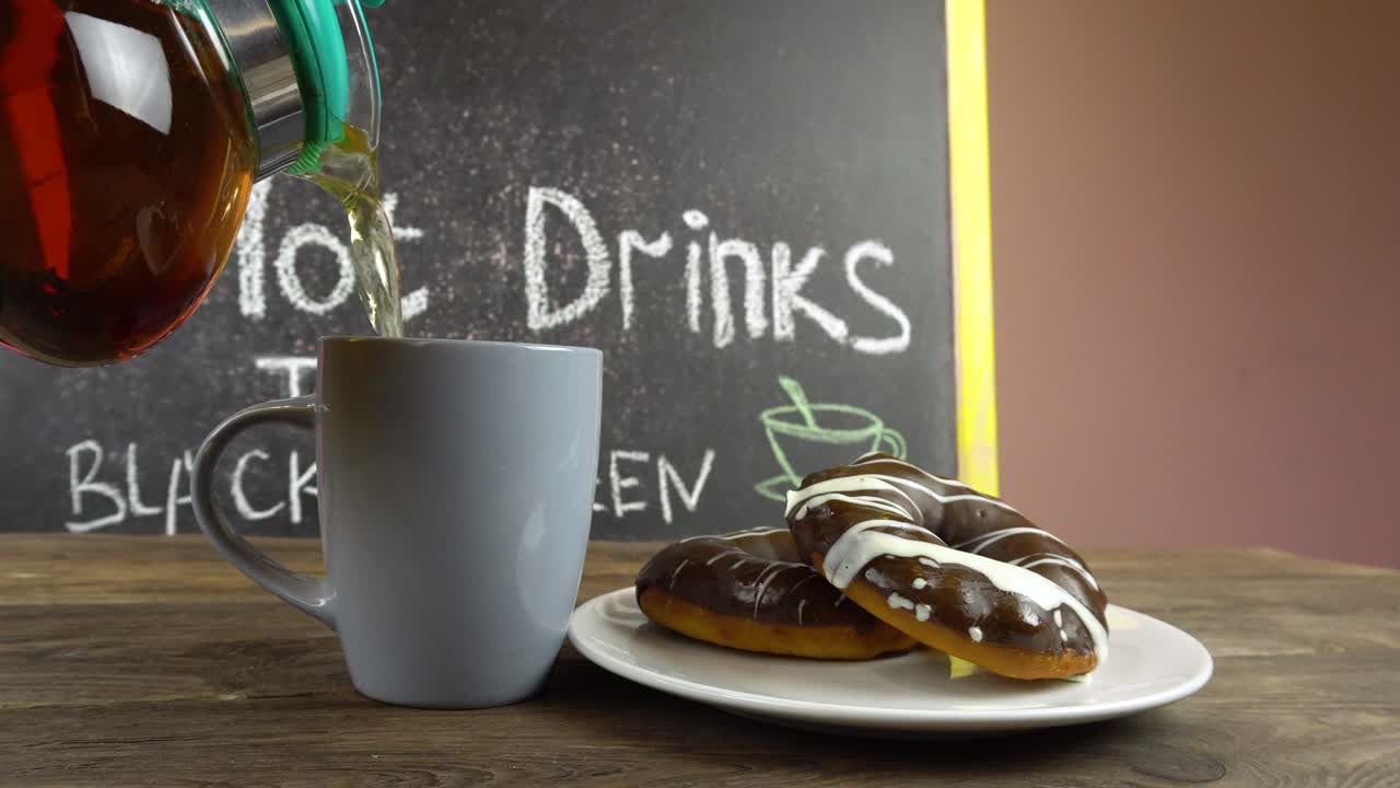 una taza de té y rosquillas en glaseado de chocolate en una mesa de café. el té está derramando de una tetera transparente.
