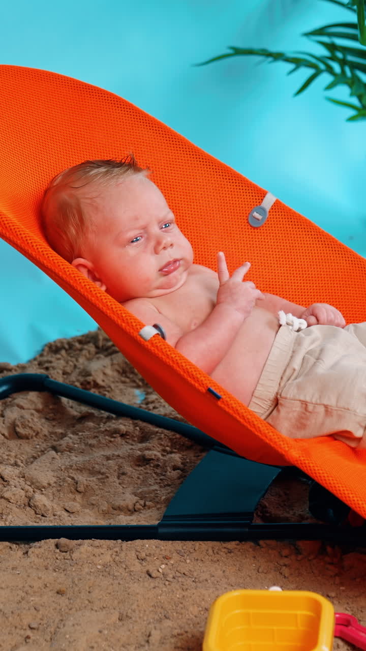 Serious newborn boy in shorts sitting in the orange baby chair. Kid resting on the improvised beach in studio. Vertical video