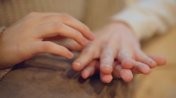 Close up of two hands gently resting and interacting on soft fabric surface with fingers overlapping in calm contact. scene features warm lighting that highlights texture of skin tones