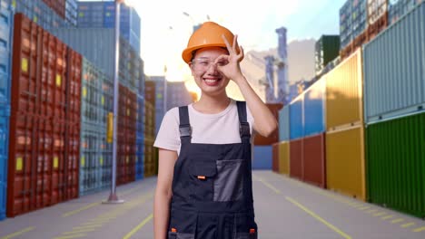 asian woman worker wearing goggles and safety helmet smiling and showing okay gesture to camera while standing at container yard warehouse