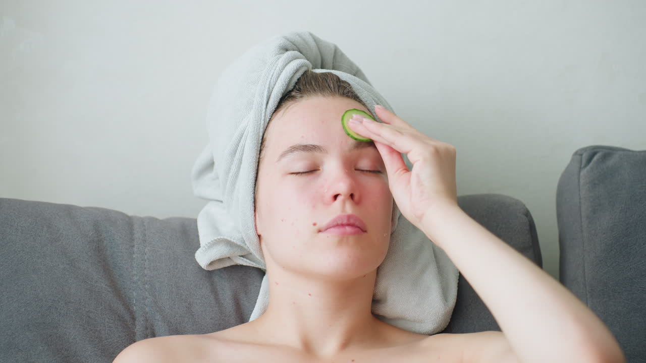 Close-up view of woman with towel-wrapped hair and green towel across chest gently rubbing cucumber slice on forehead with eyes closed, engaging in relaxing facial care inside bright peaceful indoor