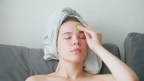 close-up view of woman with towel-wrapped hair and green towel across chest gently rubbing cucumber slice on forehead with eyes closed, engaging in relaxing facial care inside bright peaceful indoor