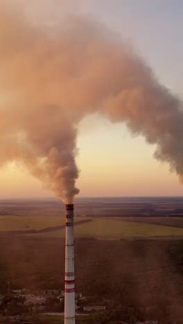 Industrial pipes with smoke. Metallurgical factory among green fields and forests in the evening. Aerial view. Vertical video