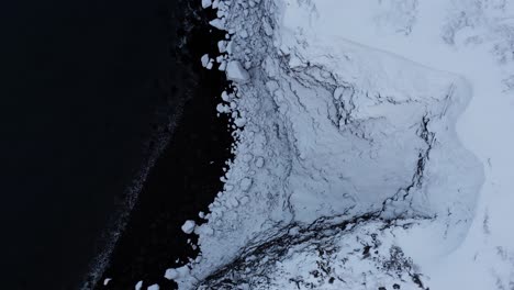 snowy coastal cliff in iceland with black volcanic sand beach, top down aerial