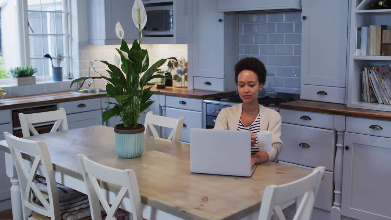 Mixed race woman using laptop and drinking coffee in kitchen