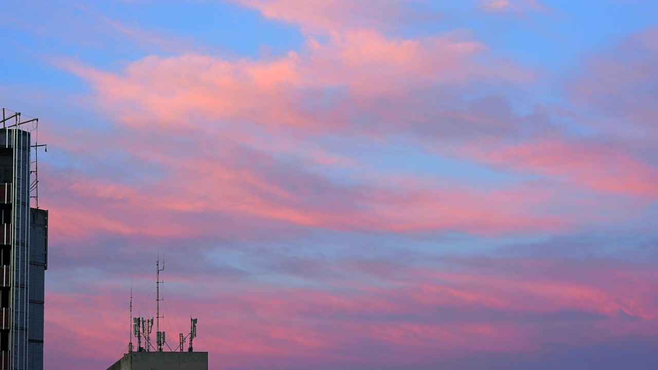 Nuvens suaves rosa e azuis espalhando-se pelo céu noturno acima de edifícios urbanos e antenas de comunicação.