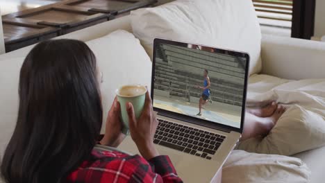 compuesto de una mujer sentada en casa sosteniendo café viendo un evento de salto de longitud de atletismo en una computadora portátil