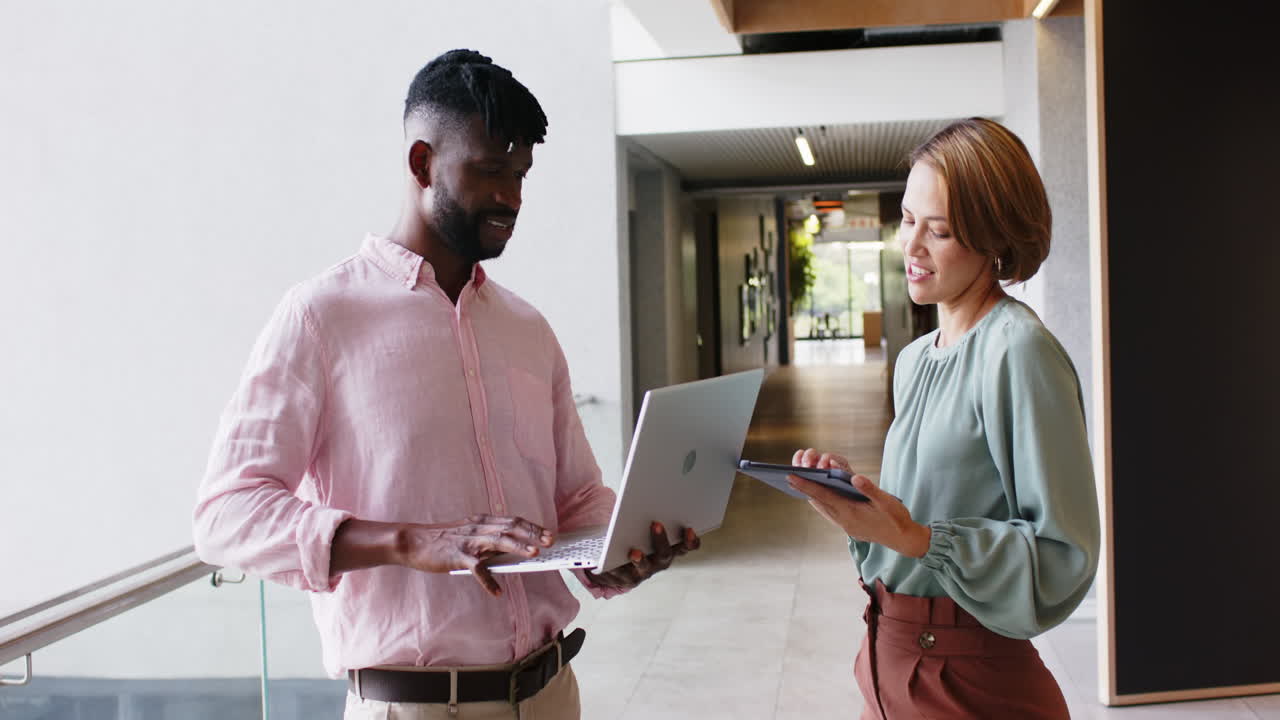 Discussing project details, colleagues using laptop and tablet in office hallway