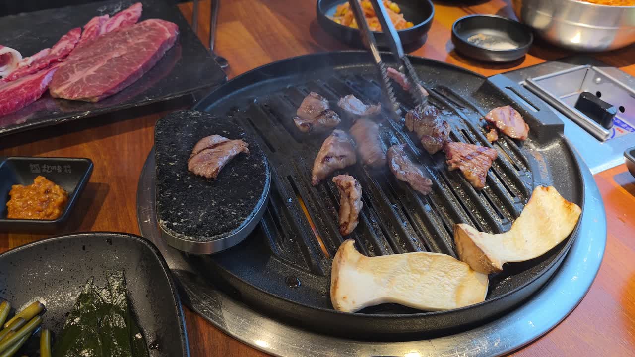 Tongs turn sizzling marbled beef and king oyster mushrooms on a tabletop grill surrounded by Korean side dishes
