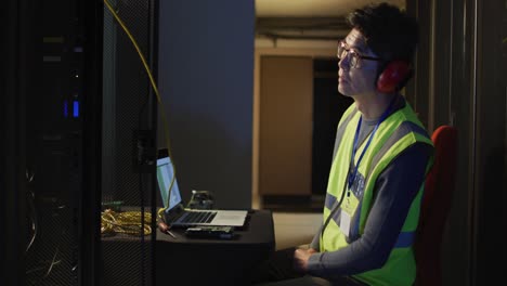 Asian-male-it-technician-wearing-headphones-and-using-torch-checking-computer-server