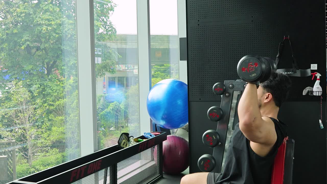 Person performing seated dumbbell shoulder press in modern Bangkok gym with fitness equipment