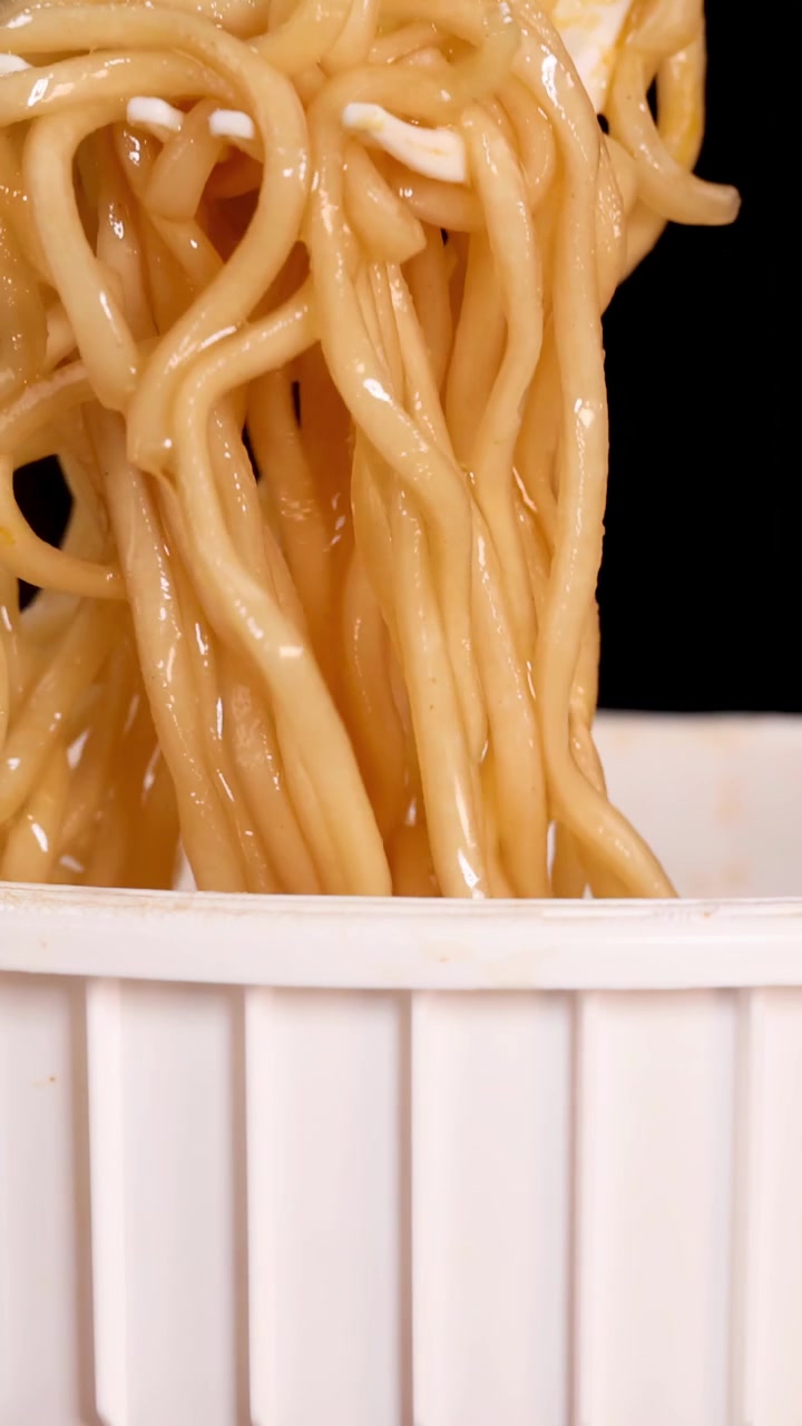 Una mano usa le bacchette per sollevare noodles istantanei cotti da una tazza bianca su uno sfondo nero, con inquadratura ravvicinata e illuminazione brillante e uniforme.