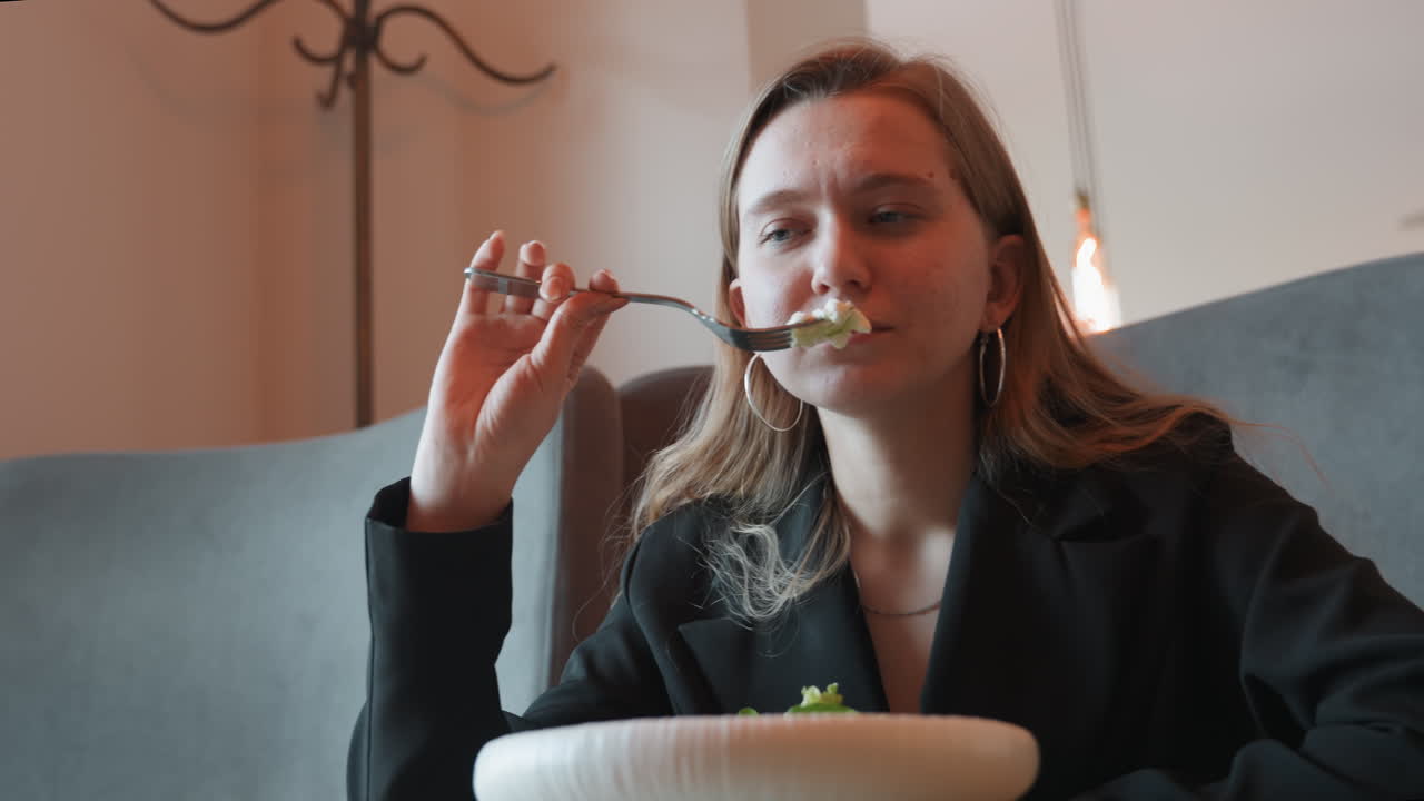 Forretningskvinde nyder en salatfrokost med selvtillid, iført blazer, med en stor skål på bordet, i en elegant holdning. Hun nyder maden langsomt i en moderne caféstemning med blød pendelbelysning, et afslappet professionelt øjeblik.