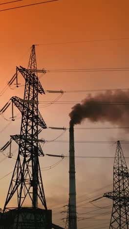 transmission lines near smoking industrial pipe at sunset. high-voltage electric power line and dark smoke released from factory in the evening. vertical video