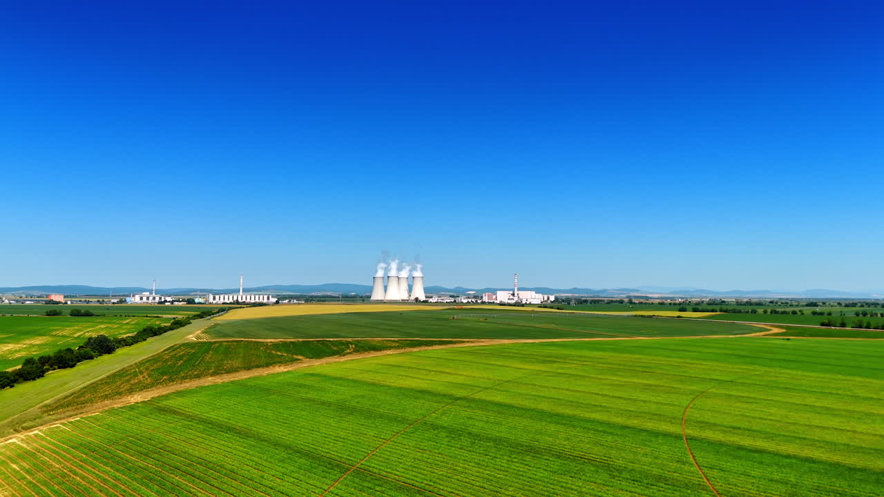 Flight above the beautiful green fields in the rural area. Four wide white tubes of the industrial plant produce thick white smoke.