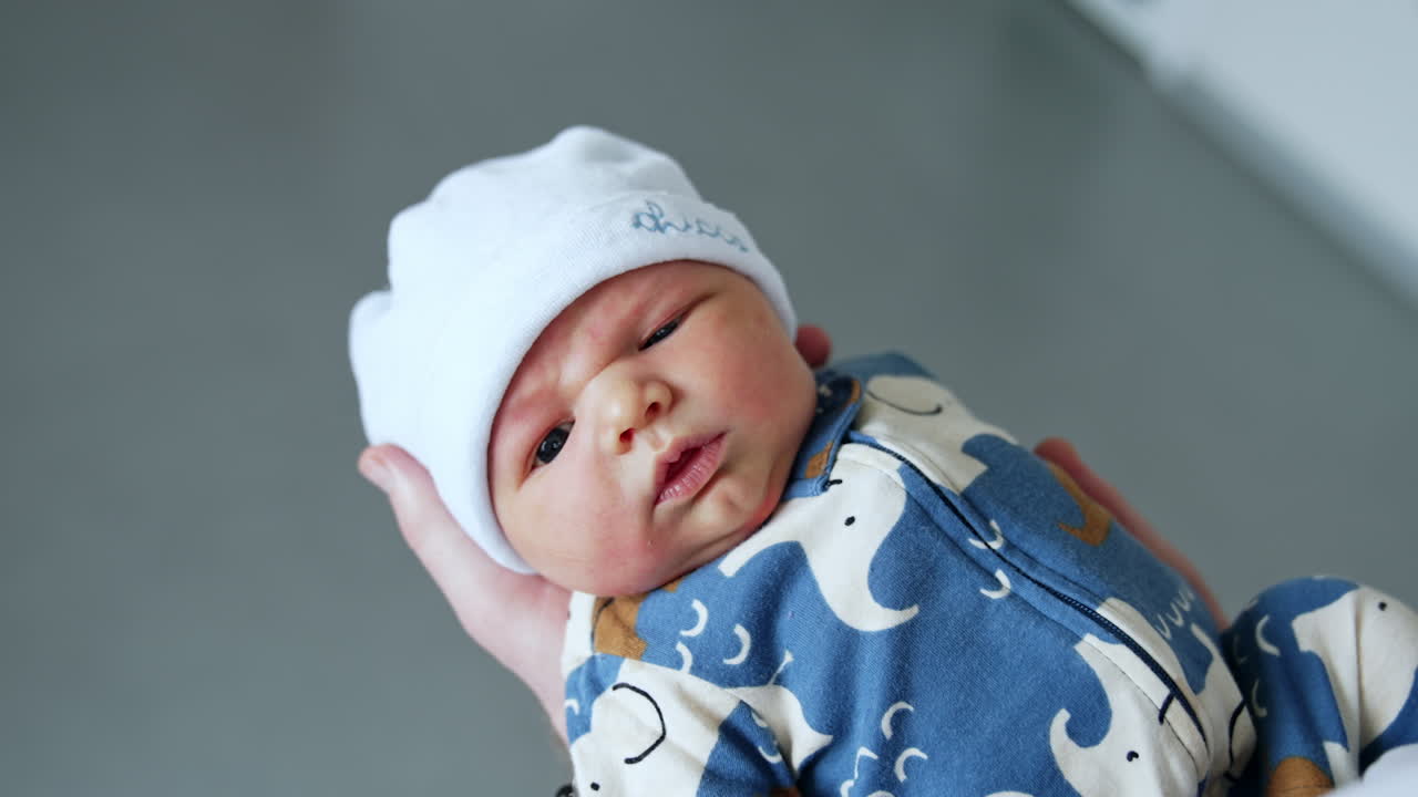 Cute dark-eyed newborn boy in blue romper and white cap. Few hours old child in dad's hands.