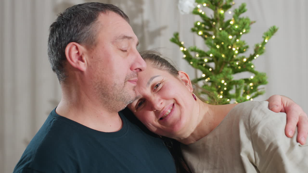 Close up view of husband and wife wearing casual clothes as woman relaxes her head on man's shoulder with peaceful smiles in cozy room decorated with Christmas tree and glowing lights