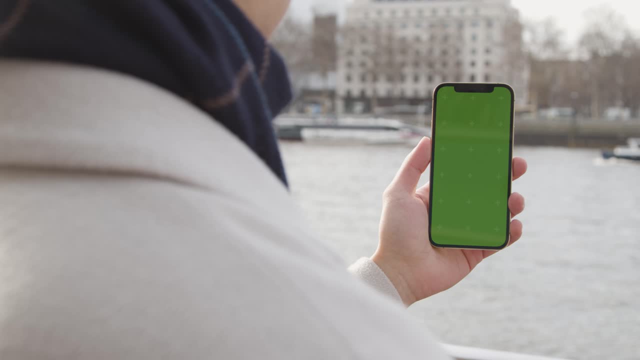primer plano de un hombre que sostiene un teléfono móvil con pantalla verde mirando hacia el río támesis y el terraplén en londres, reino unido 1