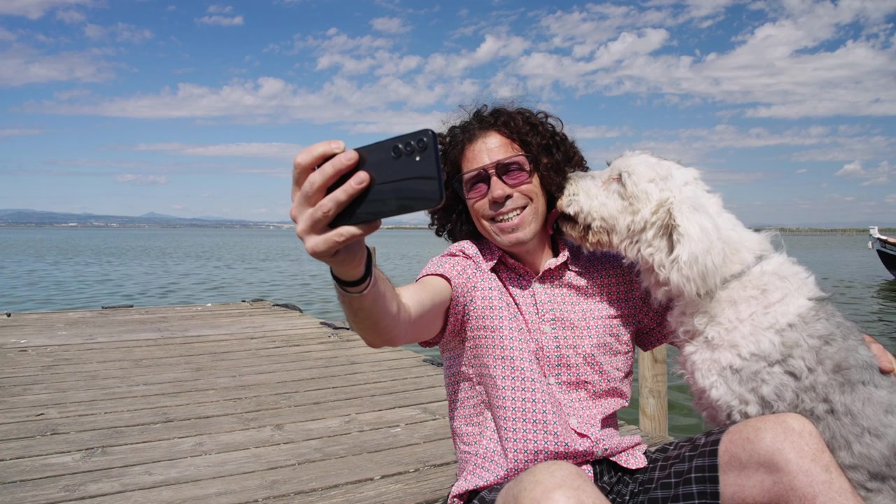 Uomo che ride mentre il suo cane gli lecca il viso mentre si scattano un selfie insieme vicino a un lago di montagna. Ripresa con telecamera orbitante.