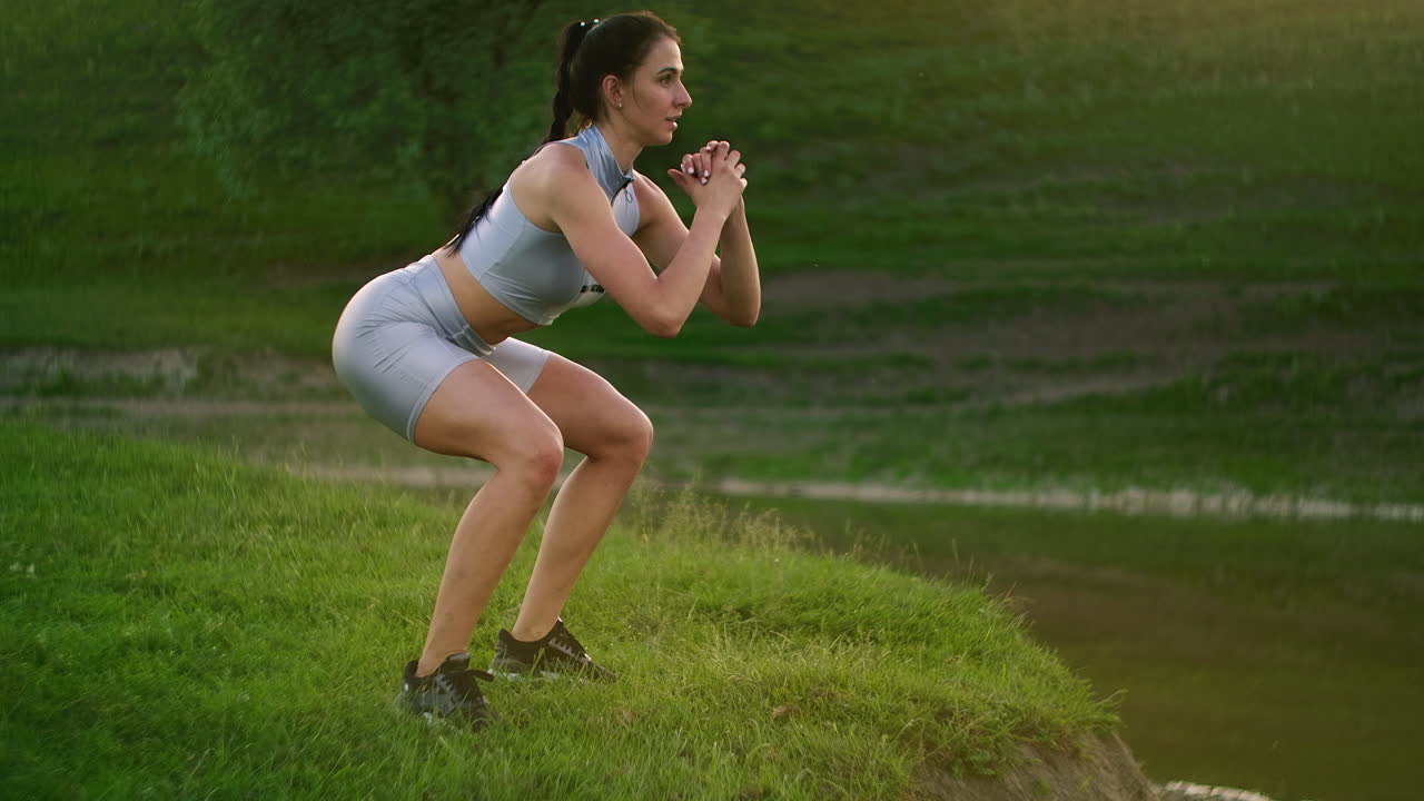 Squats and jumps up. A woman does exercises for the muscles of her thighs and legs on the grass in a Park near the lake. Slim beautiful figure. Fitness marathon. Working on your body