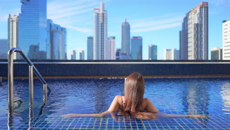 back view of woman relaxing in luxurious rooftop pool with stunning view of bangkok skyscrapers