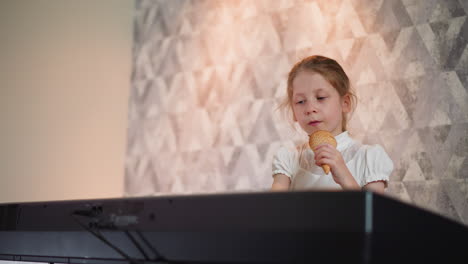 young girl wearing white top playing piano while licking ice cream cone, focused expression, warm soft lighting with patterned wall background