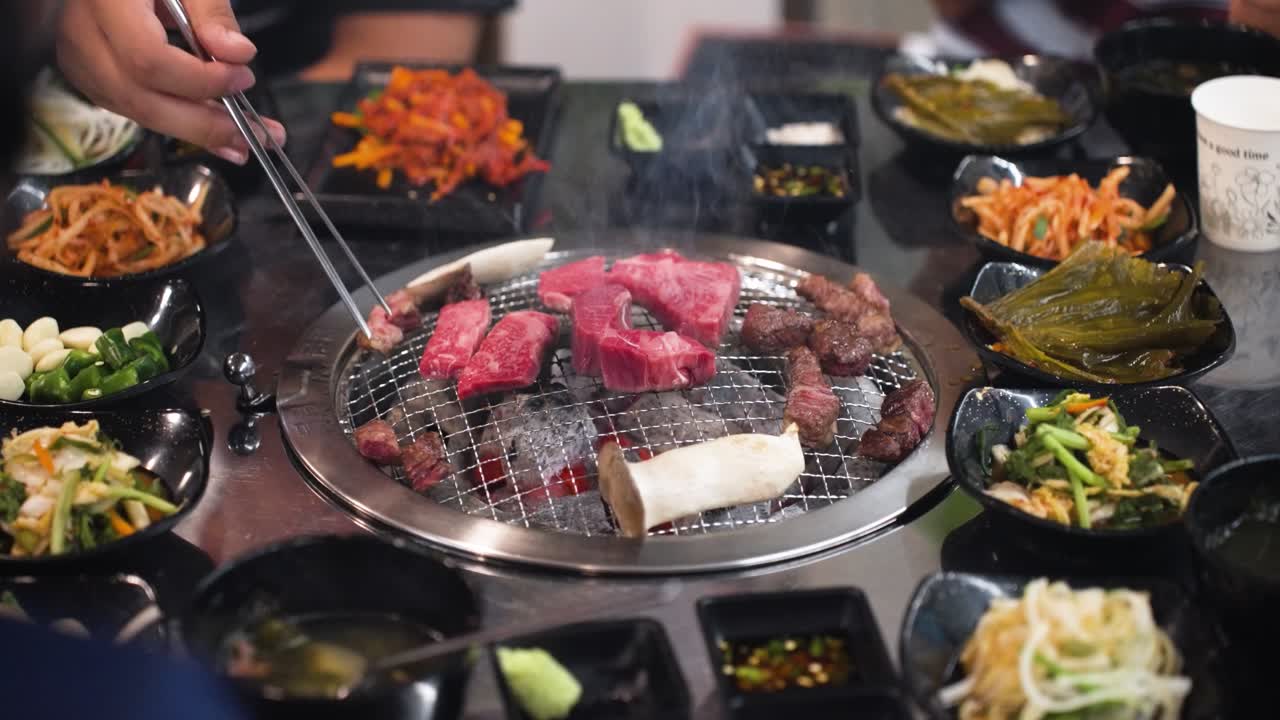 People using chopsticks and enjoying traditional Korean side dishes and noodles as slices of marbled beef sizzle on a round grill in the center of the table
