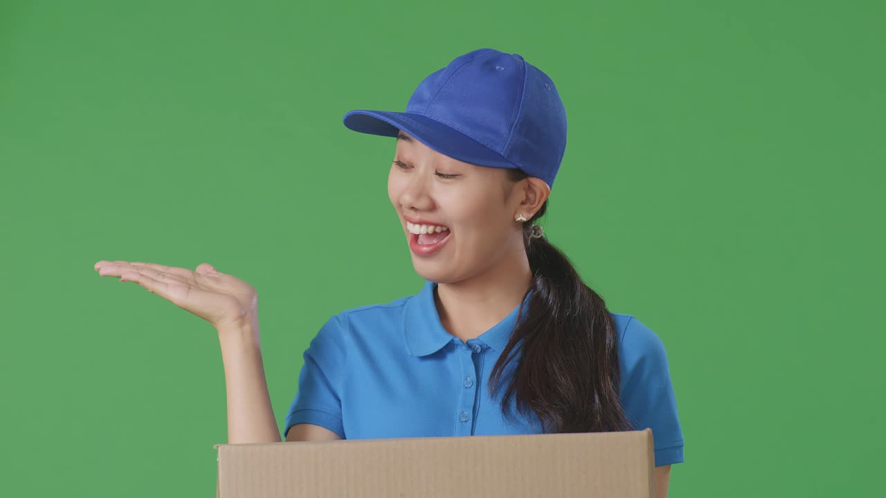 푸른 유니폼을 입은 아시아 여성 택배기사의 근접 사진. 미소 짓고 으로 가리키며 스튜디오에서 녹색 스크린 배경에 상자를 배달하고 있습니다.
