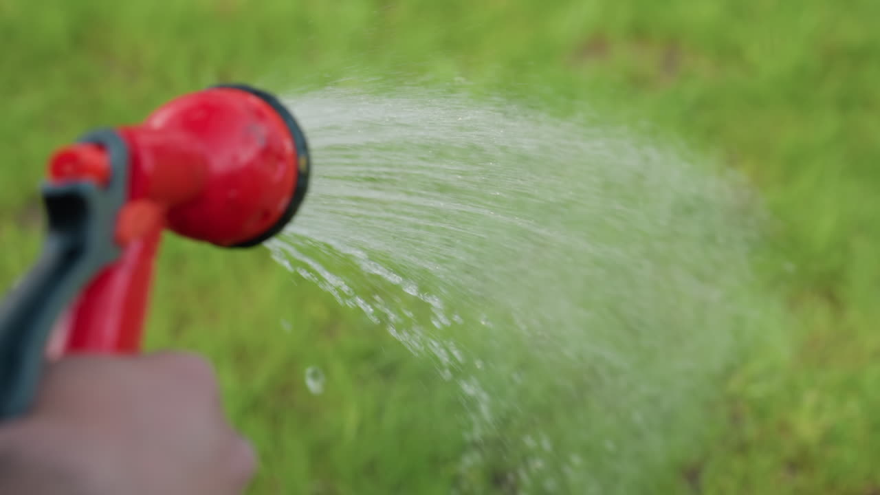 close up hand watering lush grass with garden hose, droplets sparkling on blades in crisp blur background under bright sunlight, capturing nurturing outdoor care scene of summer hydration ritual