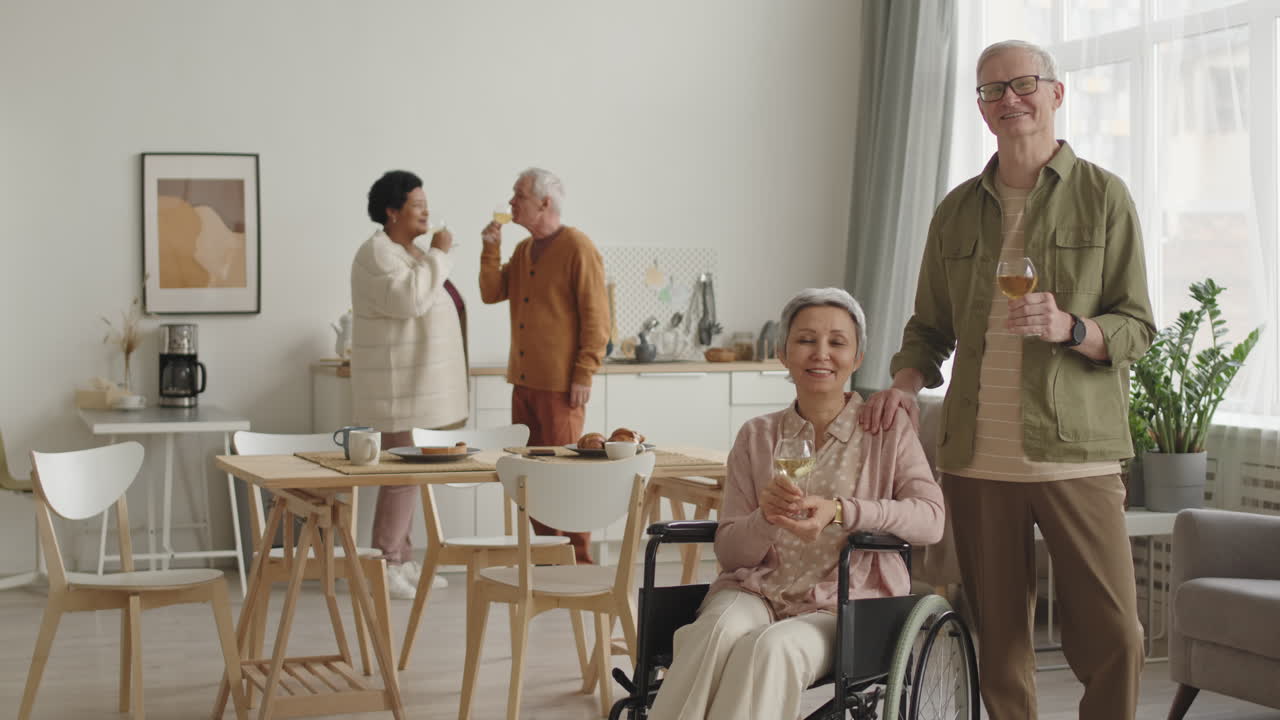 Medium long POV of silver-haired Asian handicapped woman in wheelchair smiling, clinking white wine glasses with senior Caucasian man, then spouses looking on camera, friends having fun on background