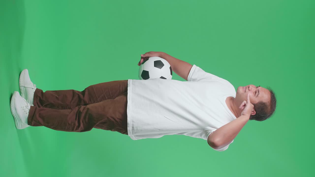 웃는 아시아 남자의 몸이 가득 차 있으며, 공을 들고 초록색 스크린 배경에서 축구를 응원하는 동안 전화 제스처를 하고 있습니다.