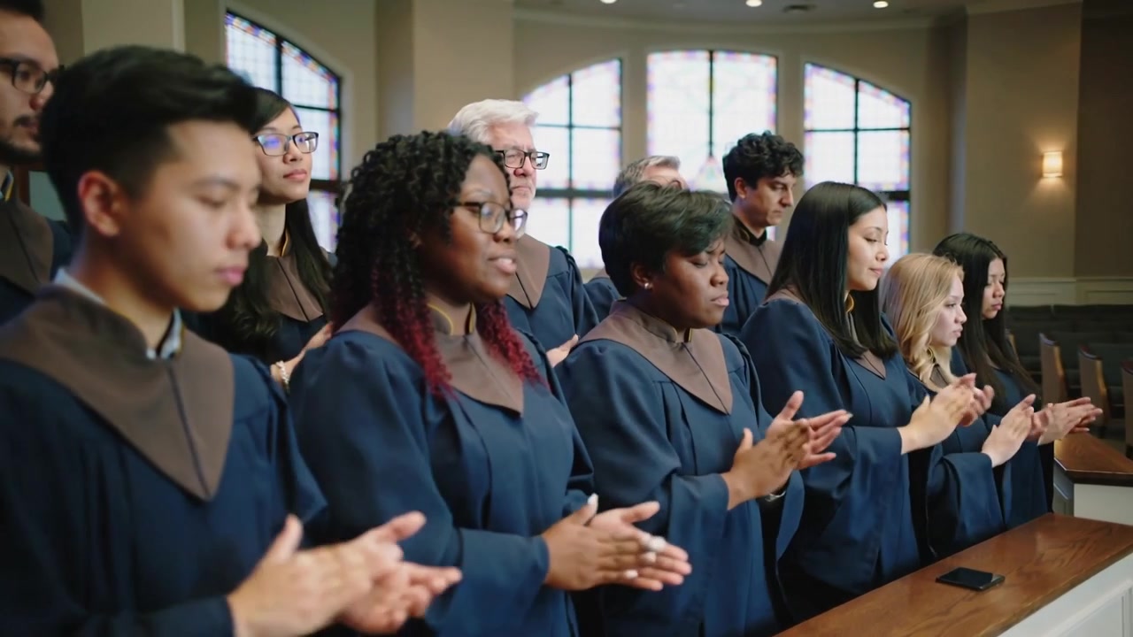 Telecamera che panoramica mostrando un coro che applaude e canta sulla balaustra della chiesa, in tonache blu scuro, per la funzione, smartphone.