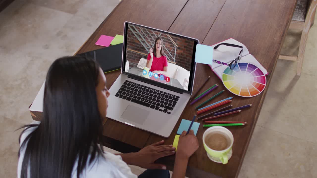 Mujer caucásica usando una laptop en videollamada con una colega, tomando notas