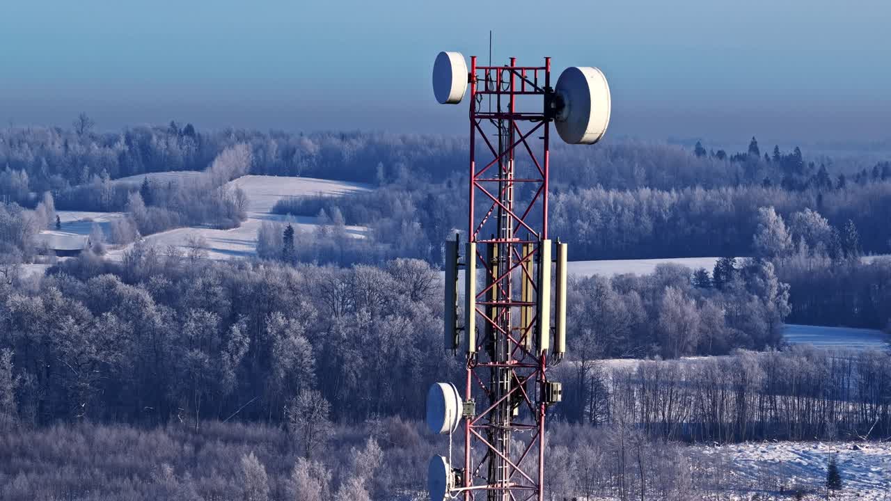 Orbiting drone shot circles red telecom tower with antennas amidst frosty birch forests rolling snowy hills under clear blue winter sky in rural Latvia