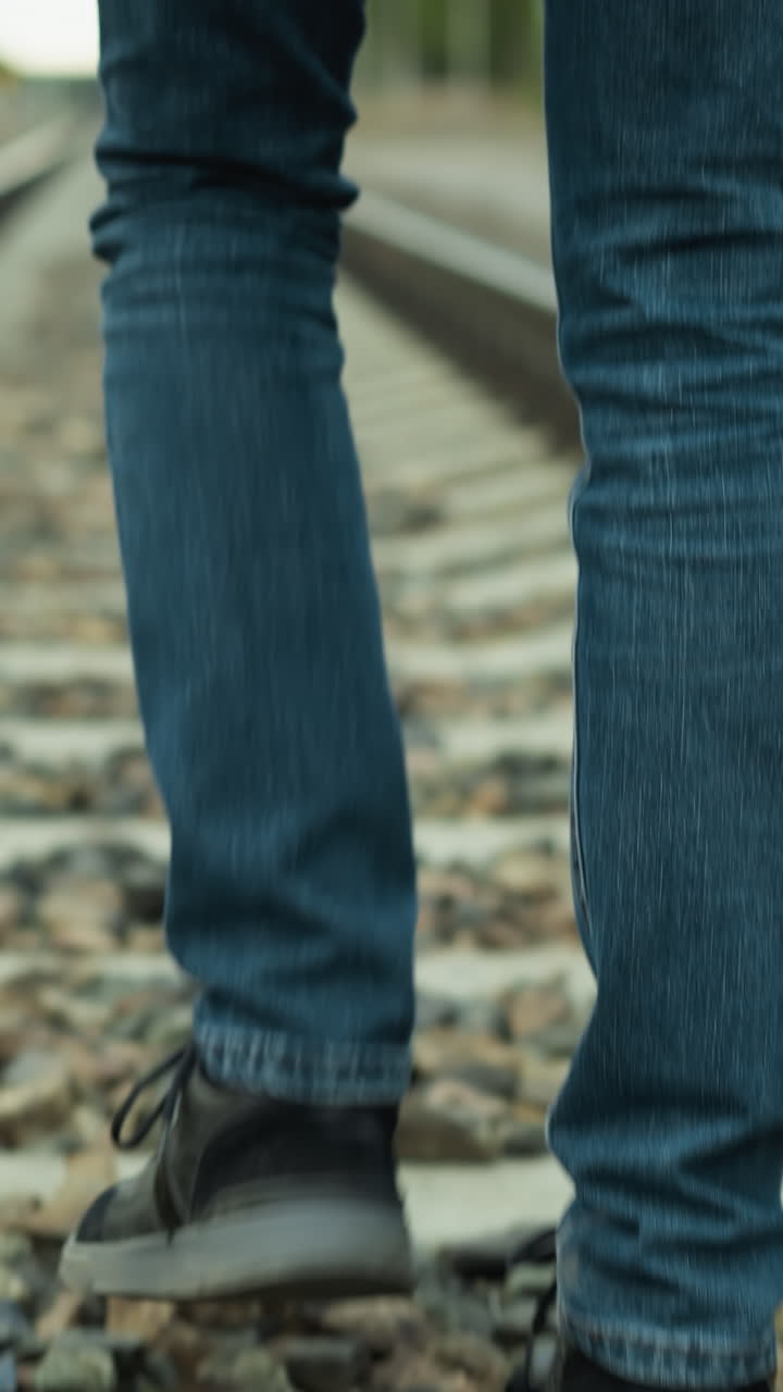 An upward view of an aged man in jeans and canvas shoes walking calmly on rocky railway tracks, with trees around and sky in view