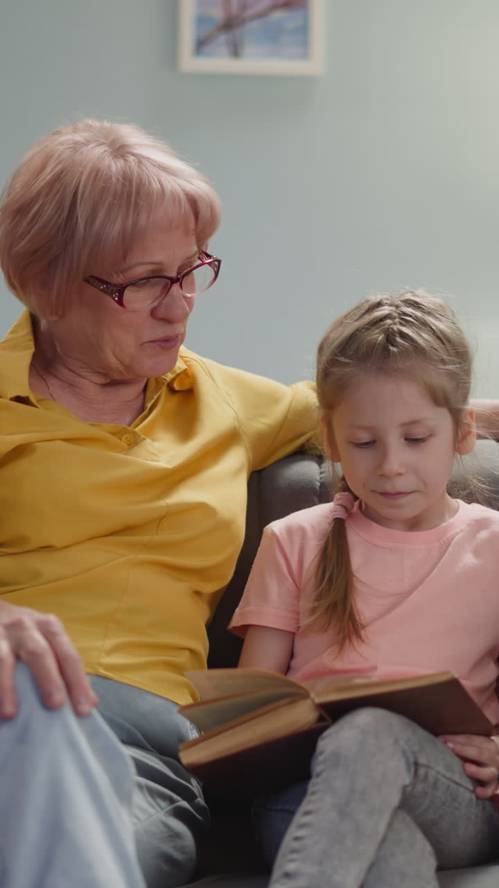 Grandmother reads book to little granddaughter resting together on sofa in living room. Mature blonde woman teaches girl to read. Family leisure