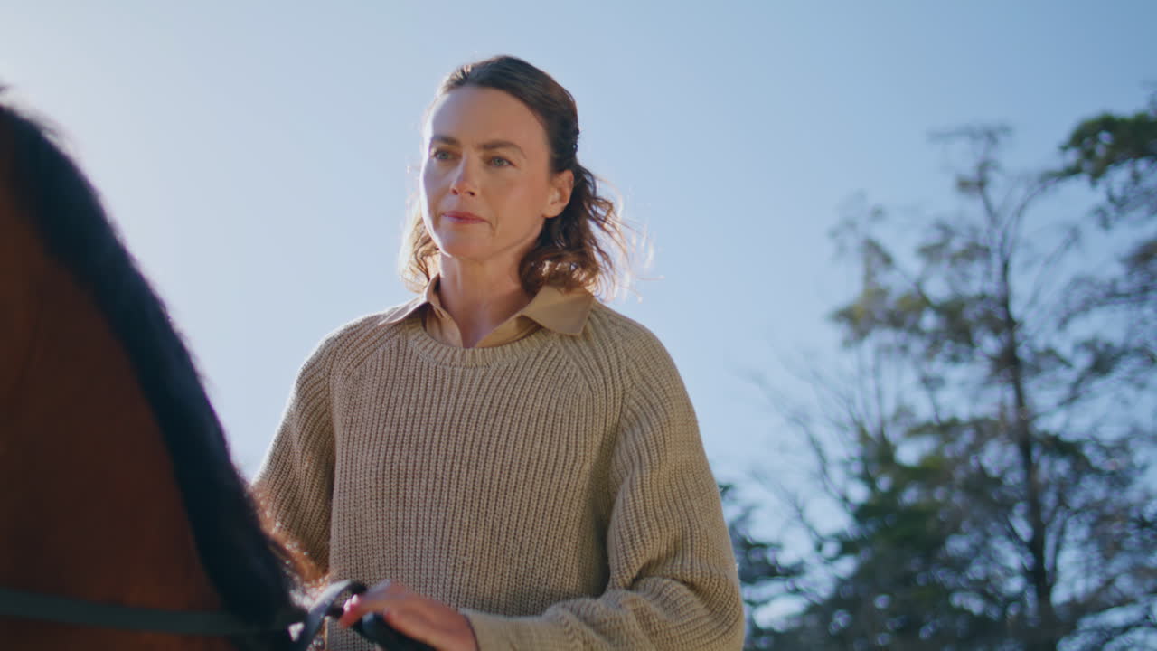Serious horsewoman holding bridle sitting stallion in saddle at nature closeup