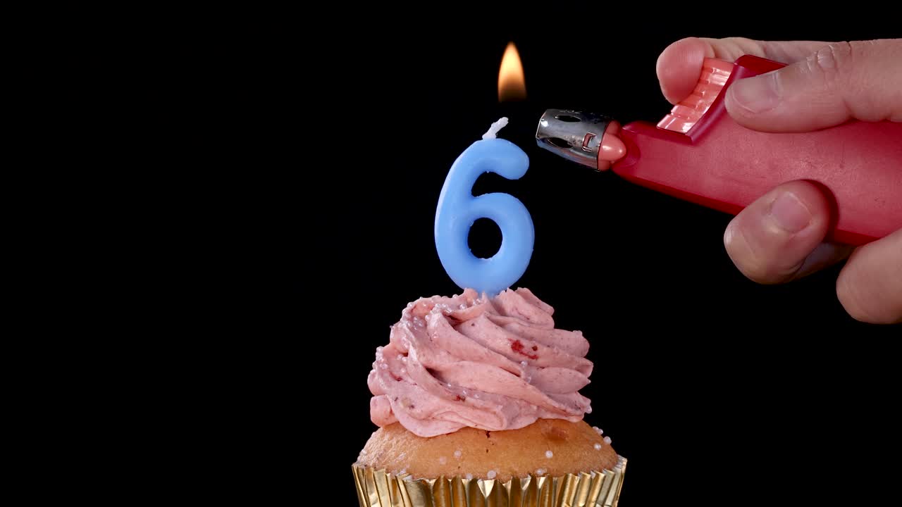 A hand lights a blue number six candle atop a strawberry frosted cupcake using a red lighter, set against a black background with steady close-up framing