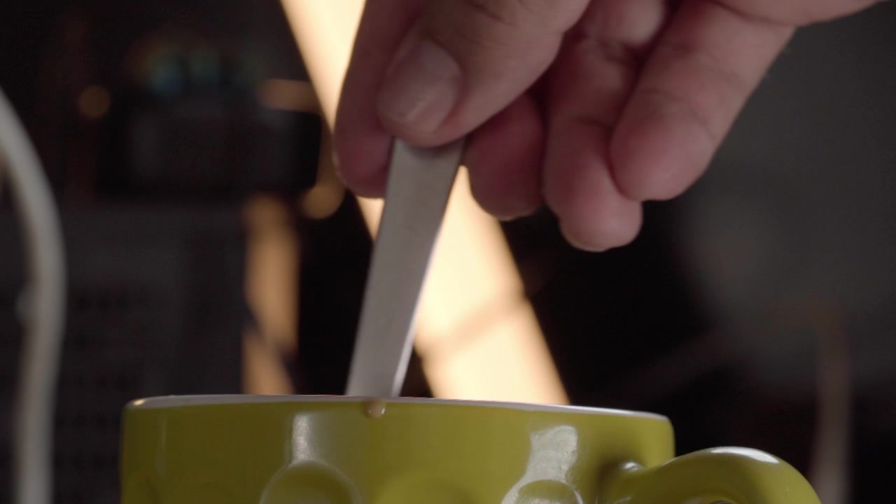 Close-up de mão mexendo café em xícara com colher durante a preparação de bebida quente. Movimento circular suave misturando a bebida naturalmente em um ambiente de cozinha acolhedor antes de servir.