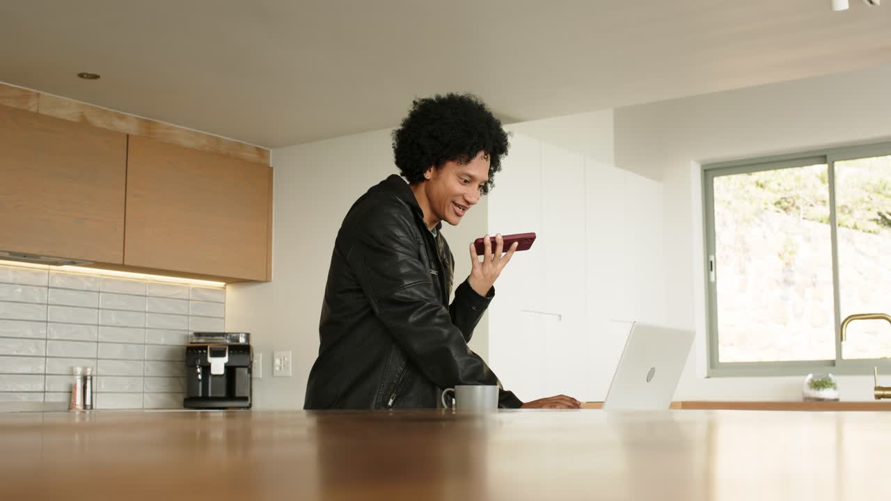Pressing smartphone to mouth, man using laptop and laughing while conversing at counter, copy space