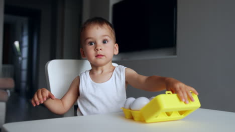 little toddler boy sits indoors at the table playing. cute kid plays with a set of white plastic eggs.